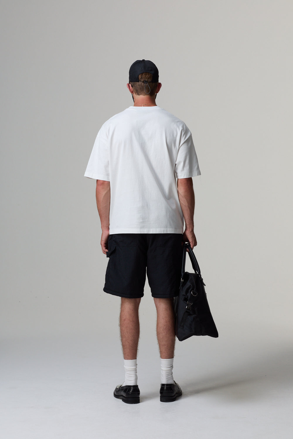 Model wearing a white Criminal Damage t-shirt, black shorts, and a black cap, holding a black bag on a grey background