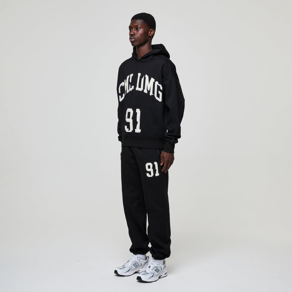 Person wearing a black hoodie and pants with text and numbers, standing against a white background