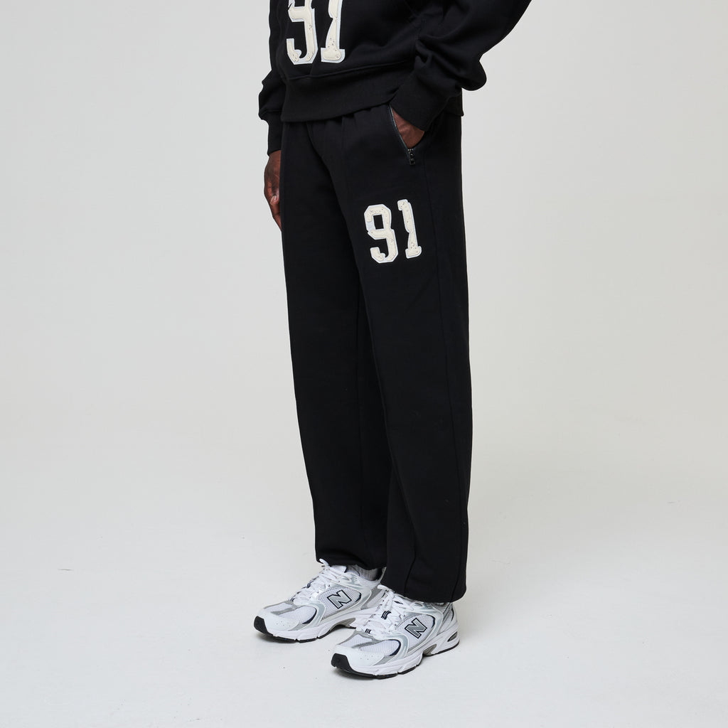 Person wearing black sweatpants with '91' printed on them, paired with white sneakers on a light gray background.