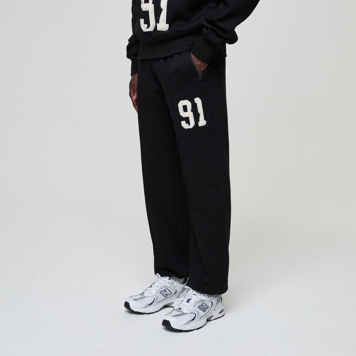 Person wearing black sweatpants with '91' printed on them, paired with white sneakers on a light gray background.