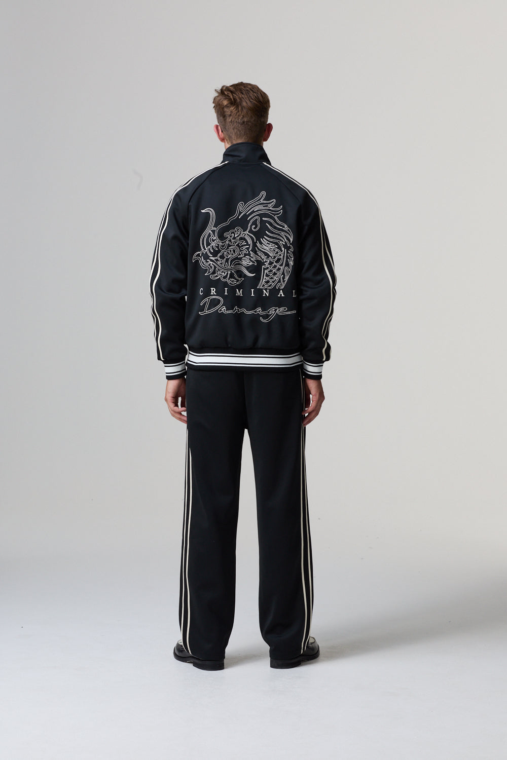 Model wearing a black Criminal Damage tracksuit with a dragon design on the back against a grey background