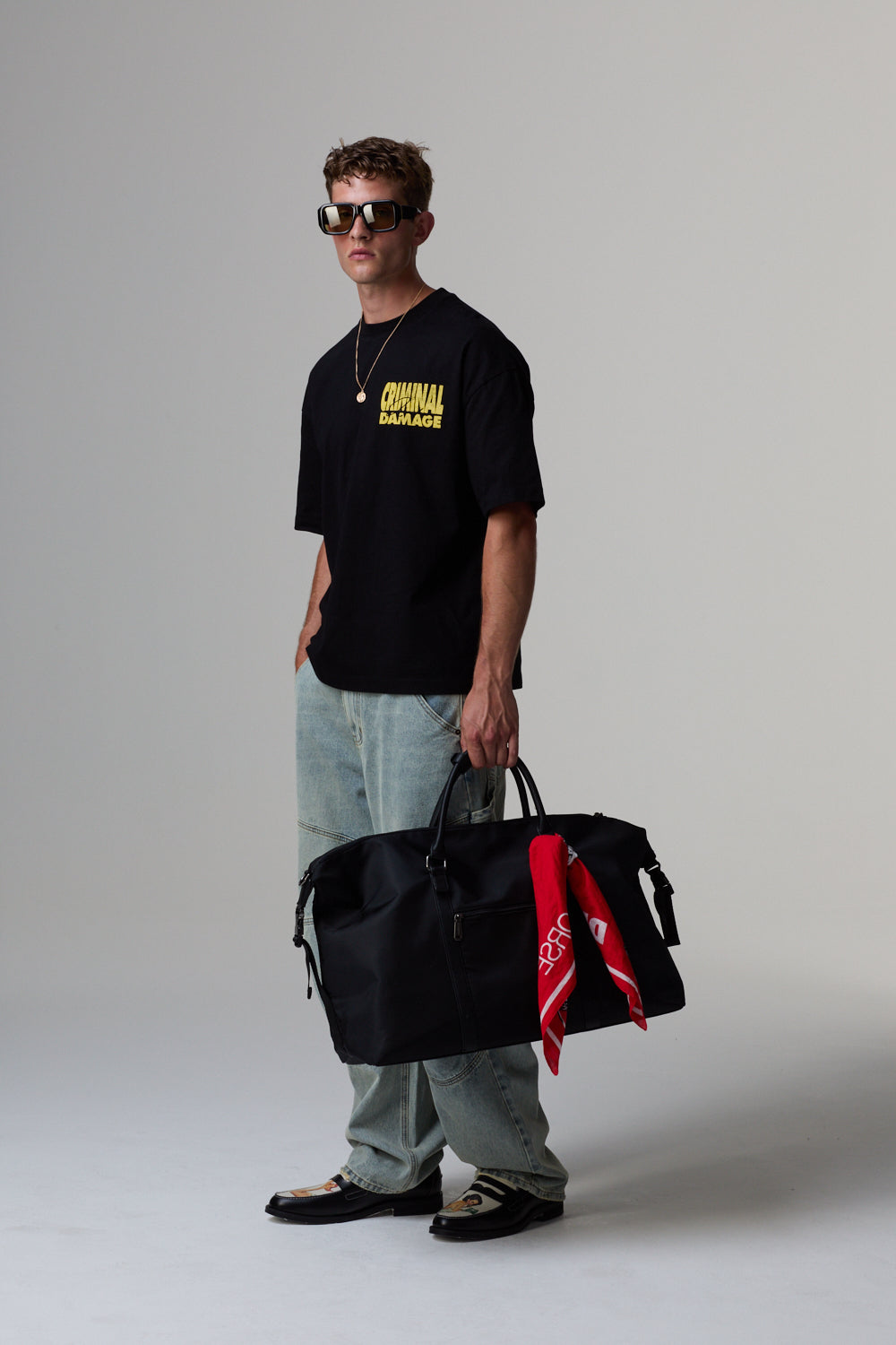 Model wearing a black Criminal Damage t-shirt with yellow text, holding a black bag with a red bandana, on a grey background.