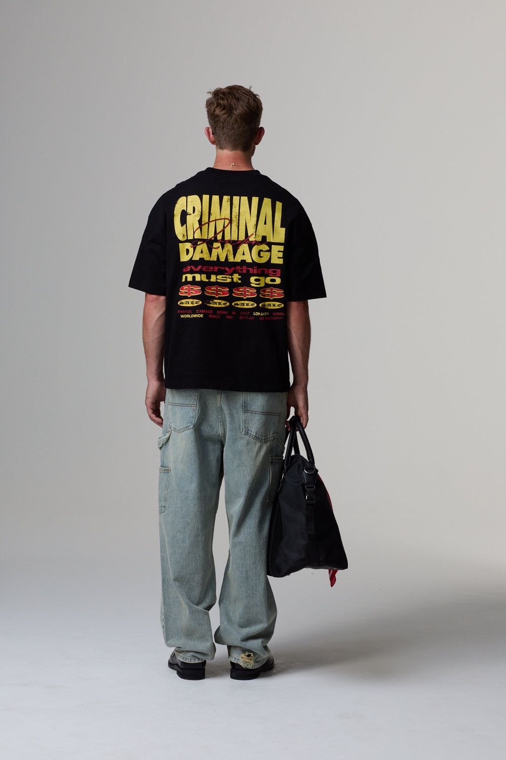 Model wearing a black 'CRIMINAL DAMAGE' t-shirt with text on the back, standing against a grey background.