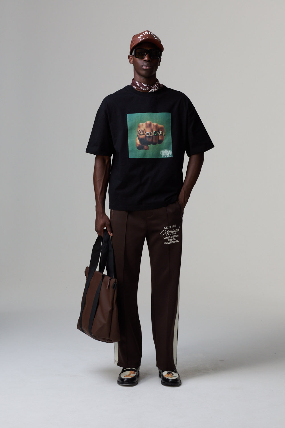 Model wearing a black Criminal Damage t-shirt with a graphic design, brown pants, and a brown cap on a grey background