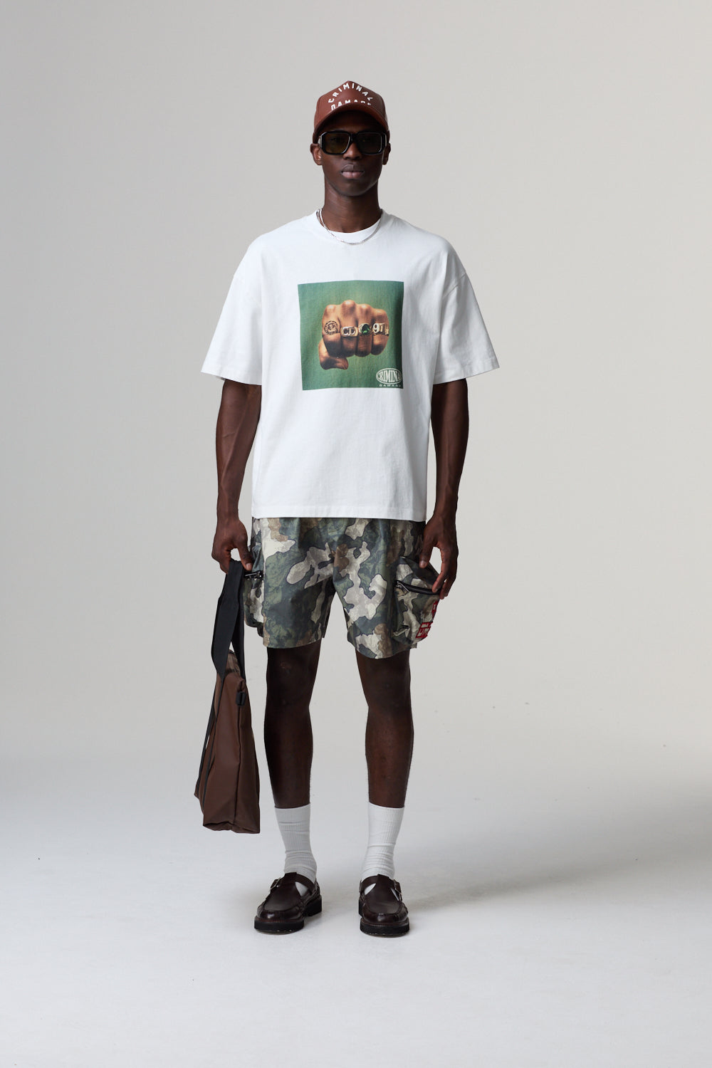 Person wearing a white t-shirt with a graphic design, camouflage shorts, and a cap on a plain background