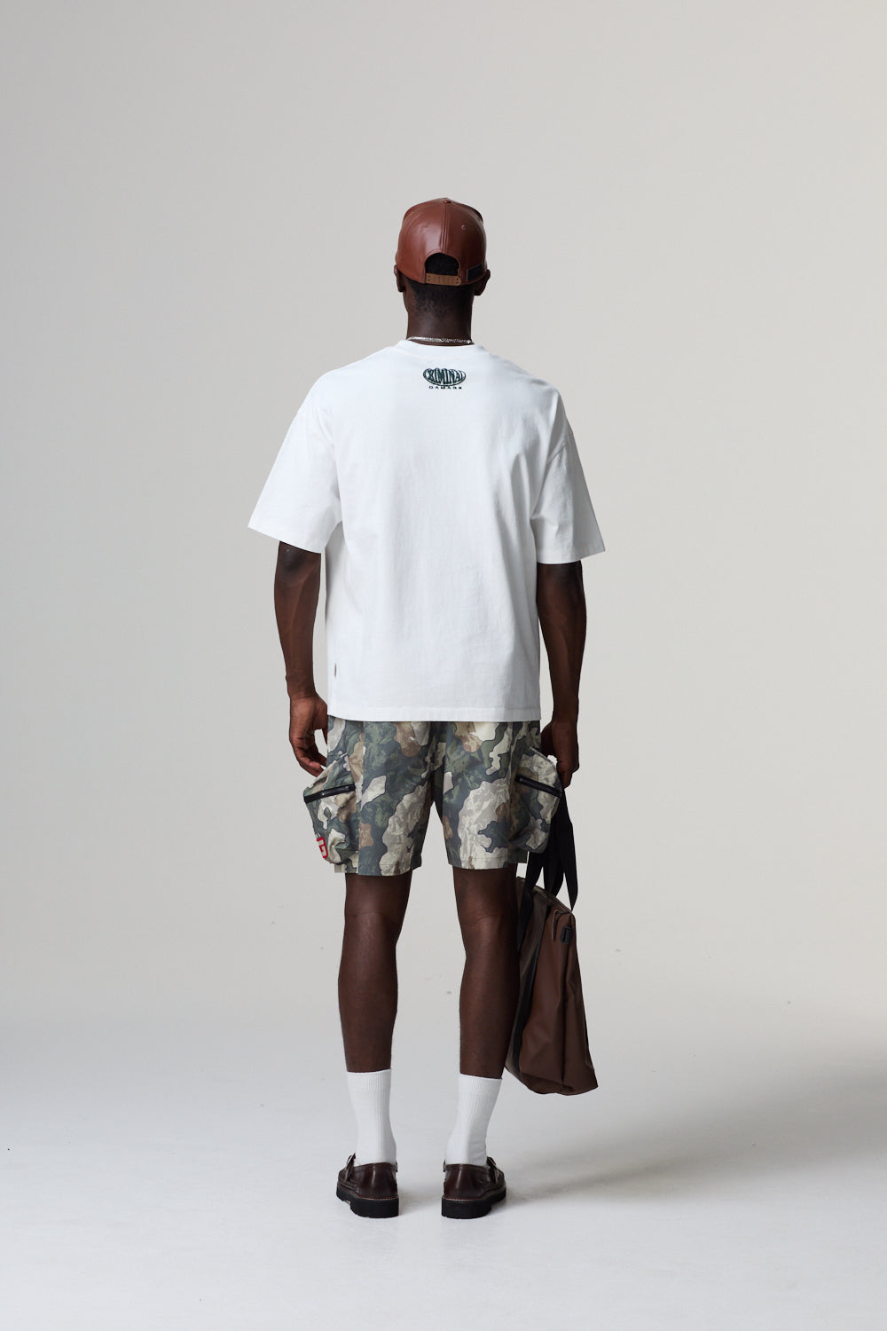 Person wearing a white t-shirt, camouflage shorts, and a red cap on a plain background