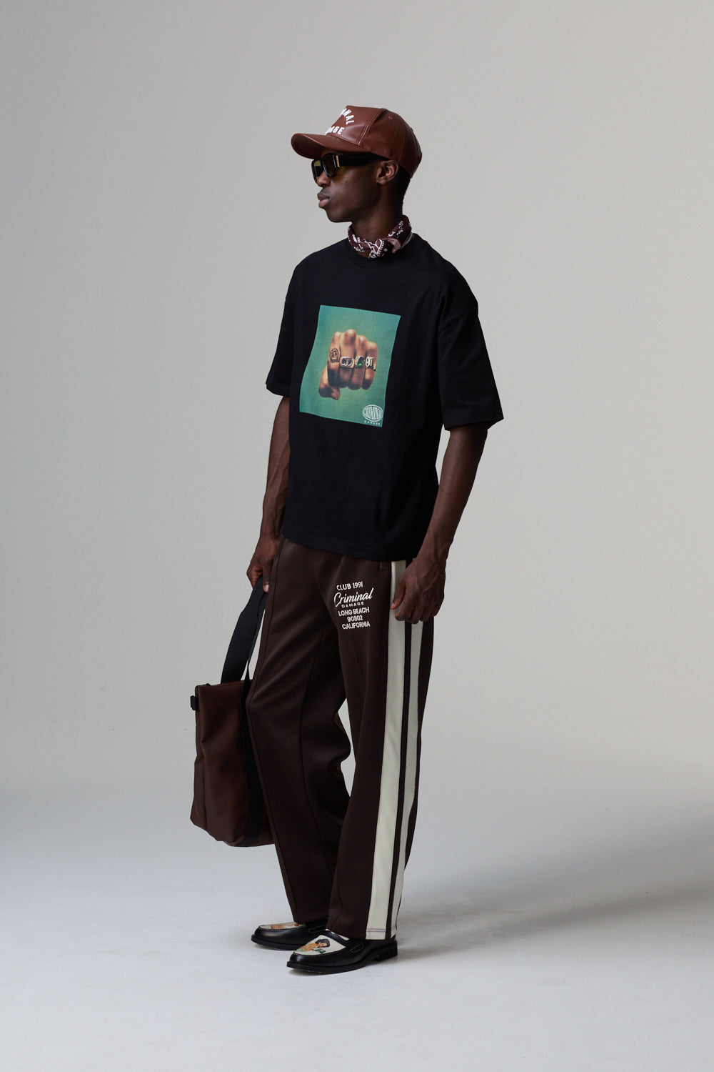Model wearing a black Criminal Damage t-shirt with a graphic design, brown pants, and a brown cap on a grey background