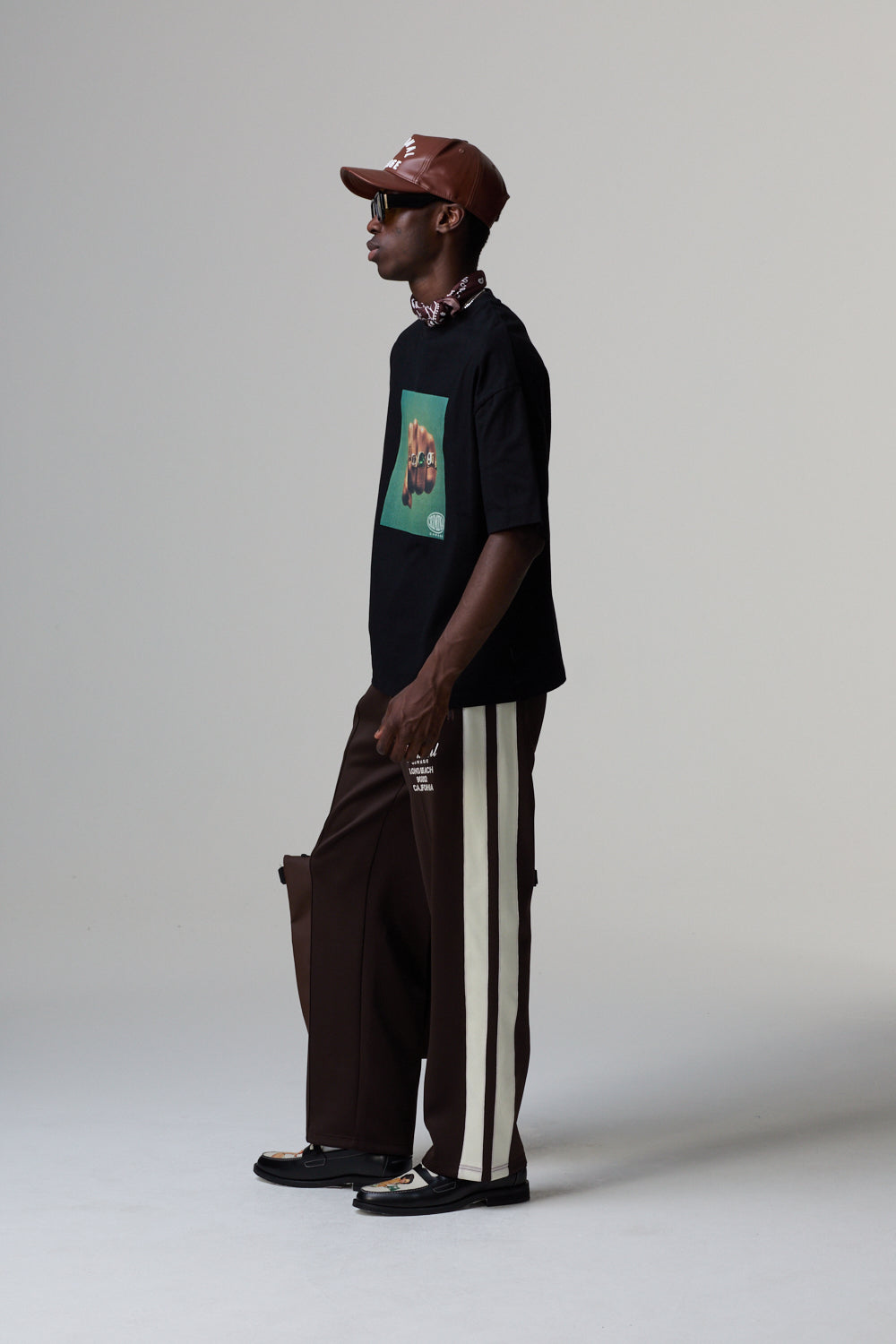 Model wearing a black Criminal Damage t-shirt with a graphic design, brown pants with white stripes, and a brown cap on a grey background.