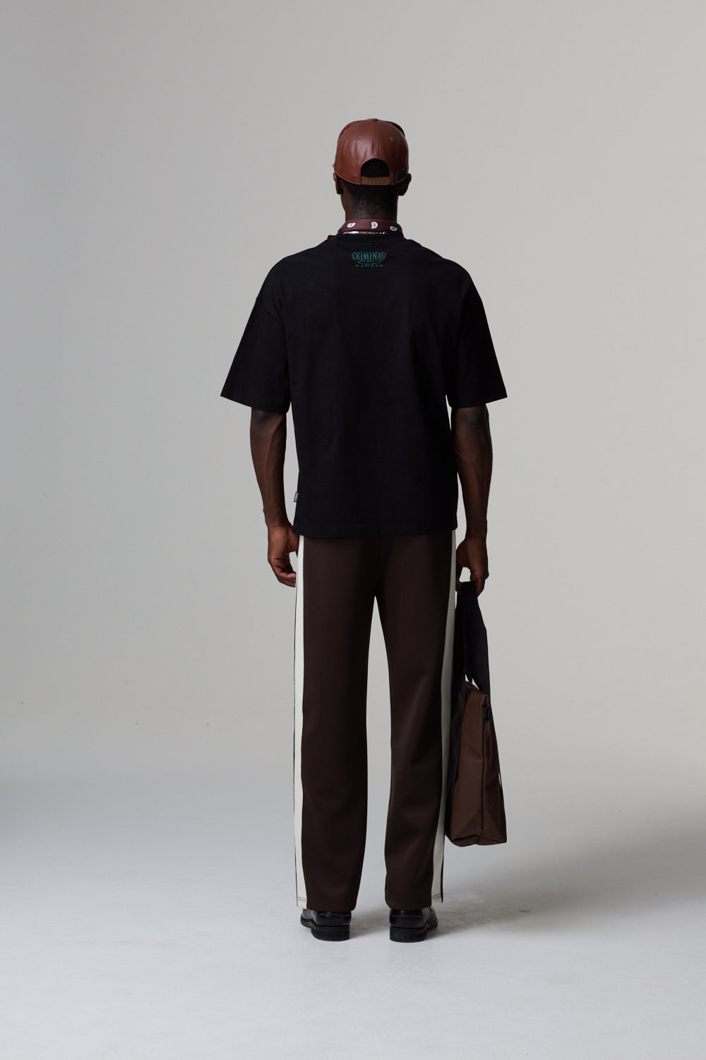Model wearing a black Criminal Damage t-shirt and brown pants holding a brown bag on a grey background
