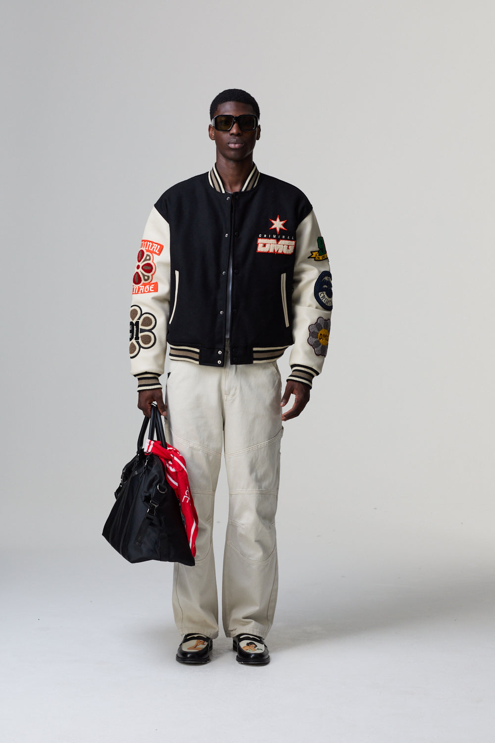 Model wearing a black and white Criminal Damage varsity jacket with colourful designs, holding a red and black bag on a grey background.