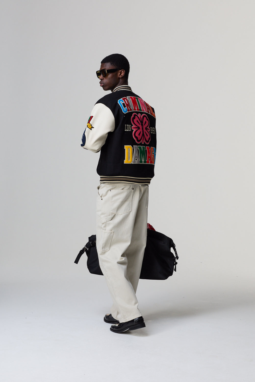 Model wearing a Criminal Damage varsity jacket with colourful text and graphics, standing against a grey background.