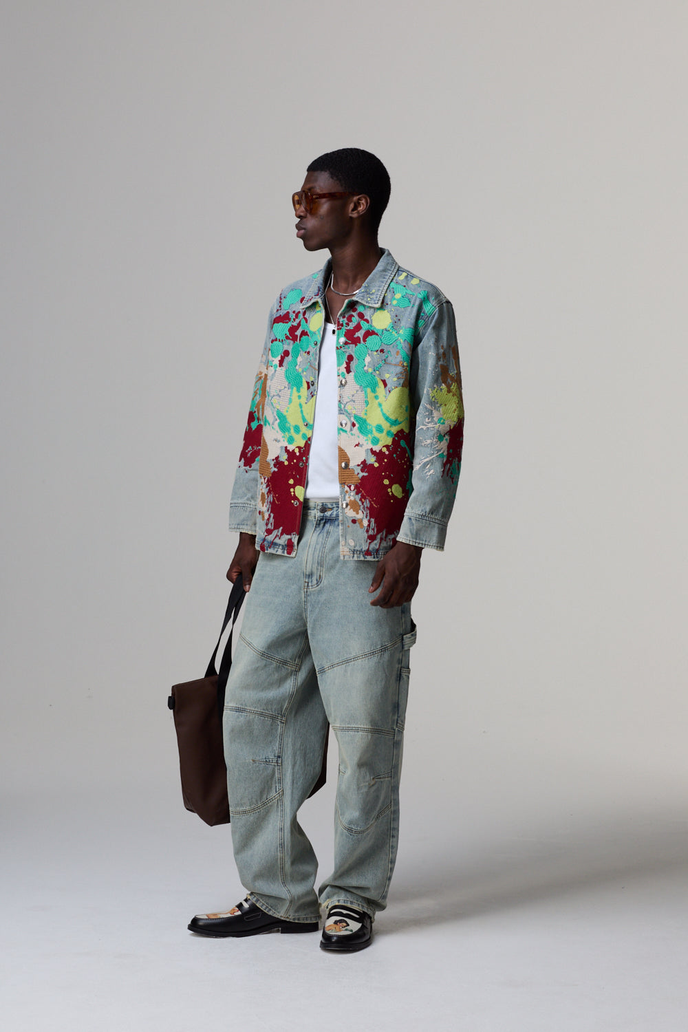 Model wearing a colourful Criminal Damage jacket and light blue jeans on a grey background