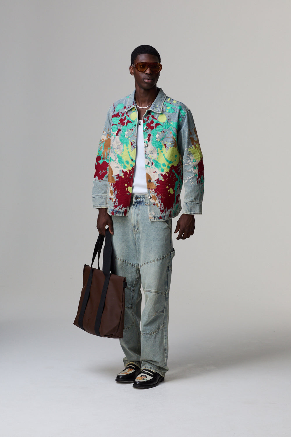 Model wearing a colourful Criminal Damage jacket and holding a brown bag on a grey background
