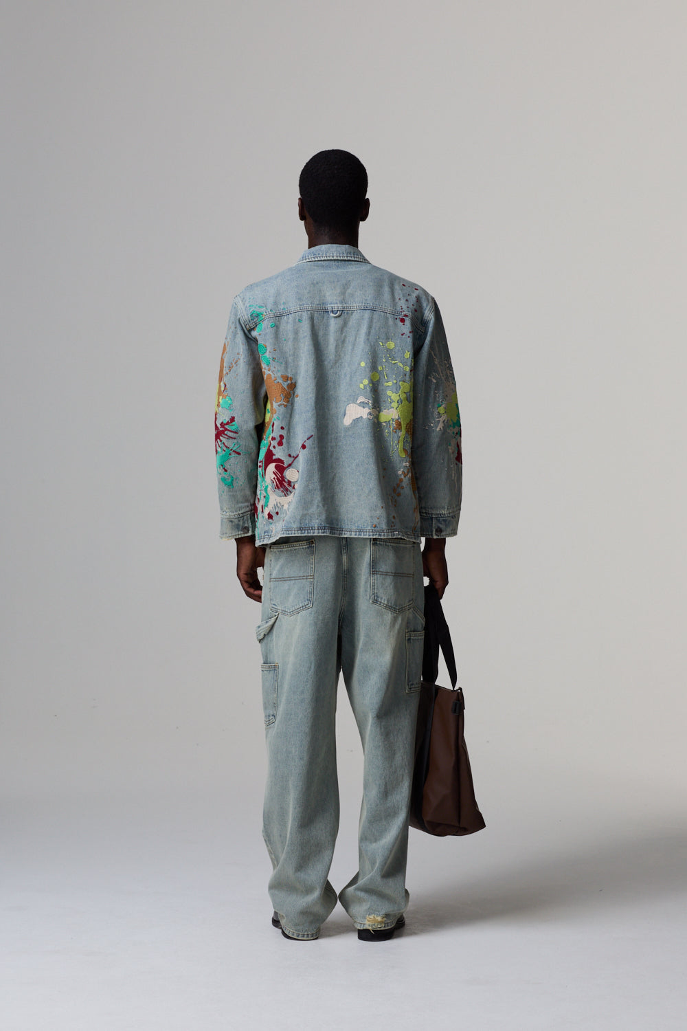 Model wearing a light blue Criminal Damage denim jacket with colourful patterns and matching pants, holding a brown bag on a grey background.