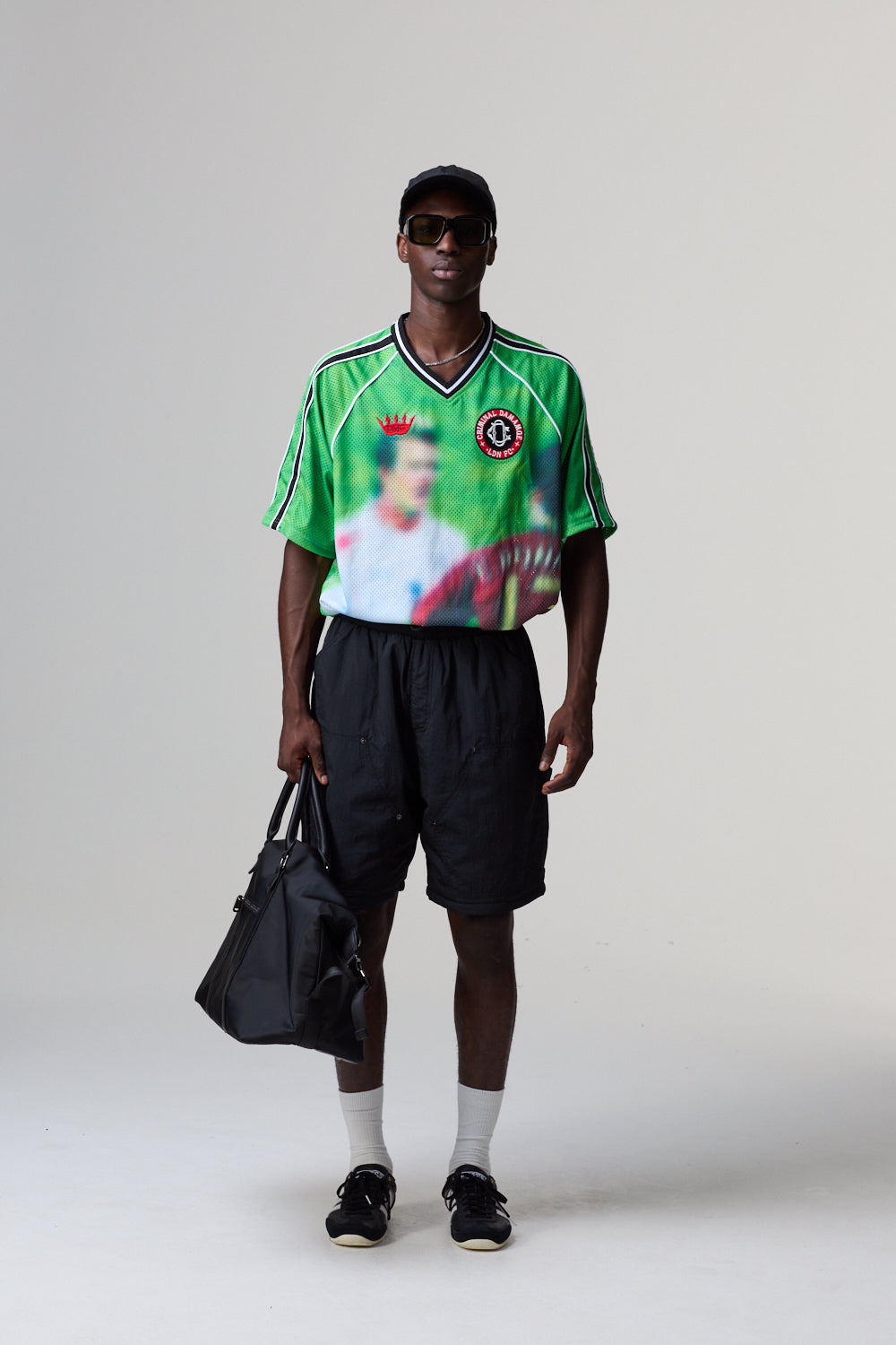 Person wearing a Criminal Damage jersey with a graphic design, holding a black bag on a grey background