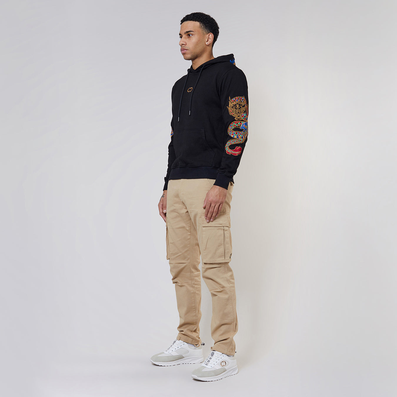 Man wearing a Criminal Damage black hoodie with a colourful design and beige cargo pants on a white background