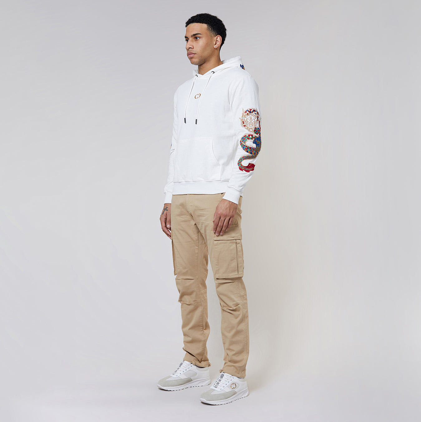 Man wearing a Criminal Damage white hoodie with colourful arm design and beige cargo pants on a gray background