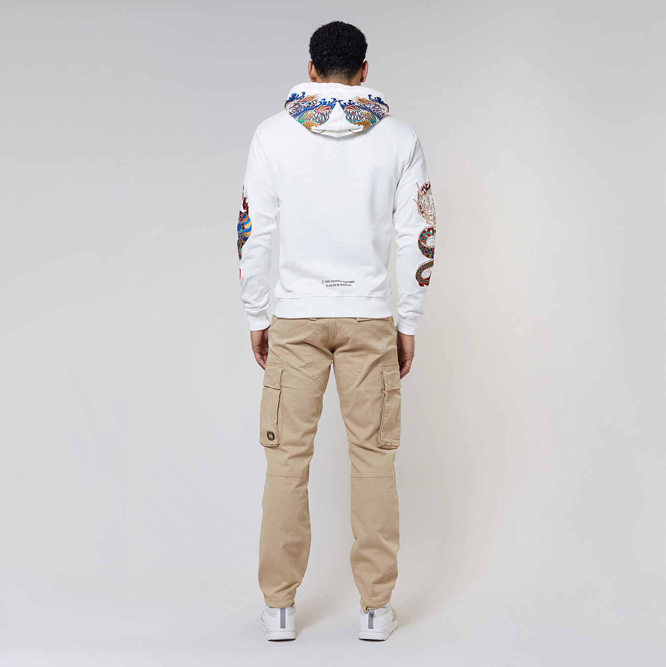 Man wearing a Criminal Damage white hoodie with colourful arm design and beige cargo pants on a gray background