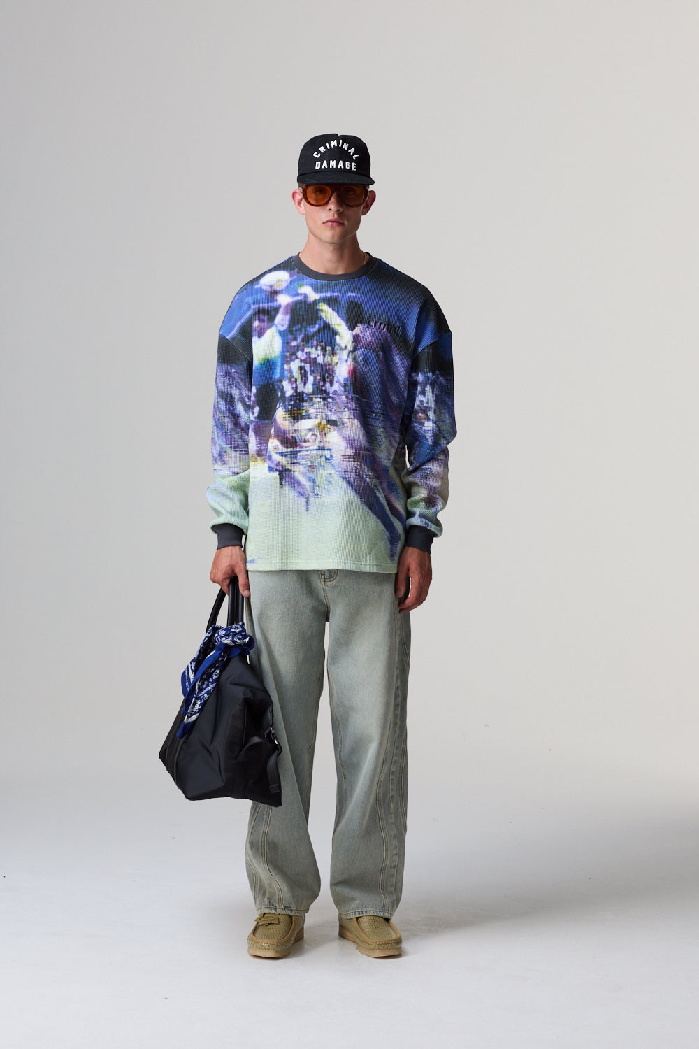 Model wearing a colourful Criminal Damage sweater, light pants, and a black bag on a plain background