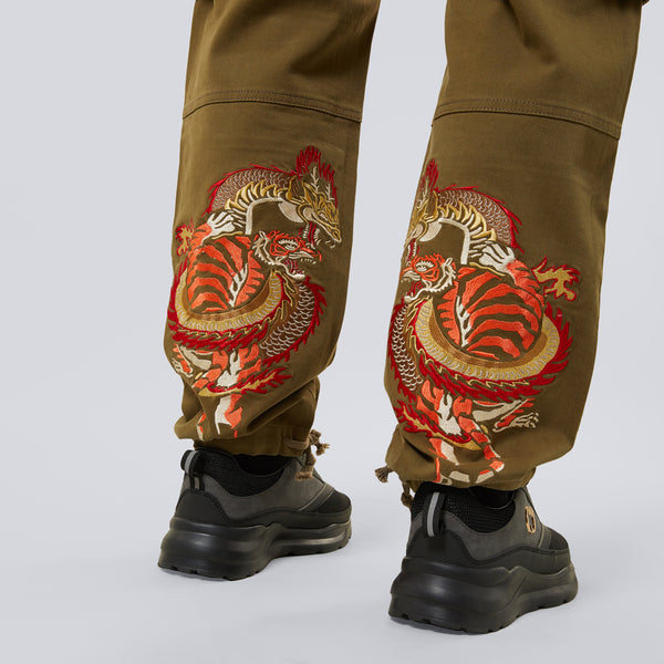 Criminal Damage brown pants with dragon embroidery worn by a person on a white background