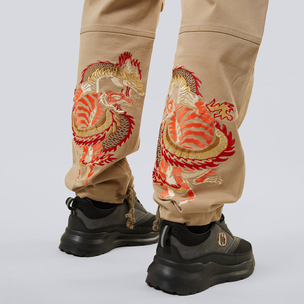 Criminal Damage beige pants with dragon embroidery worn by a person on a light grey background