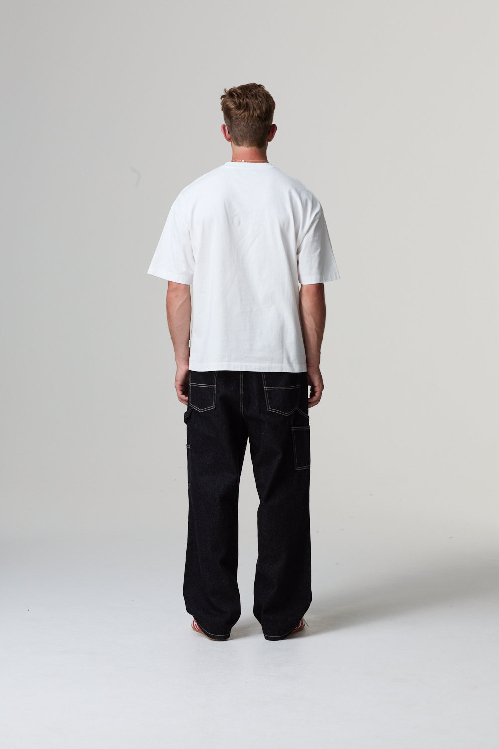 Model wearing a white Criminal Damage graphic t-shirt, red cap, and black shorts holding a black bag on a grey background