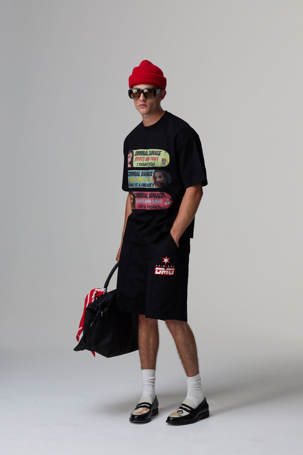 Model wearing a black t-shirt with colourful graphics, black shorts, red beanie, and holding a black bag on a grey background