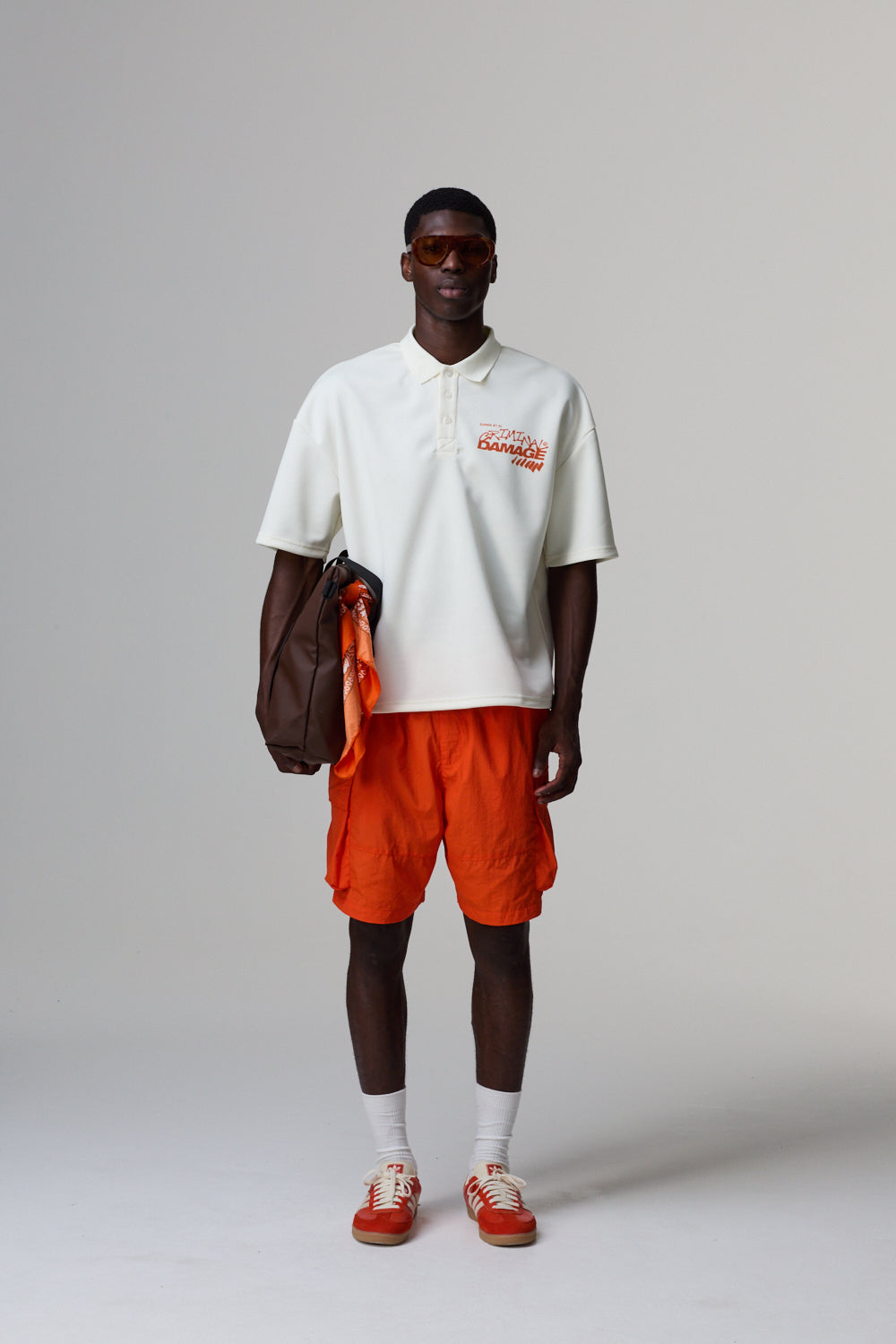 Model wearing a white Criminal Damage polo shirt with a logo, orange shorts, and red shoes on a grey background