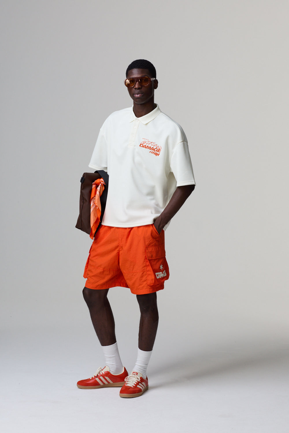Model wearing a white Criminal Damage polo shirt, orange shorts, and red shoes on a grey background