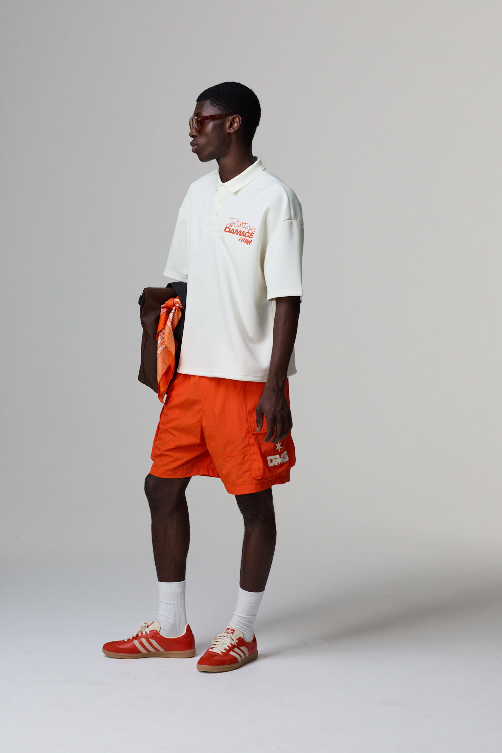 Model wearing a white Criminal Damage polo shirt, orange shorts, and red sneakers on a grey background