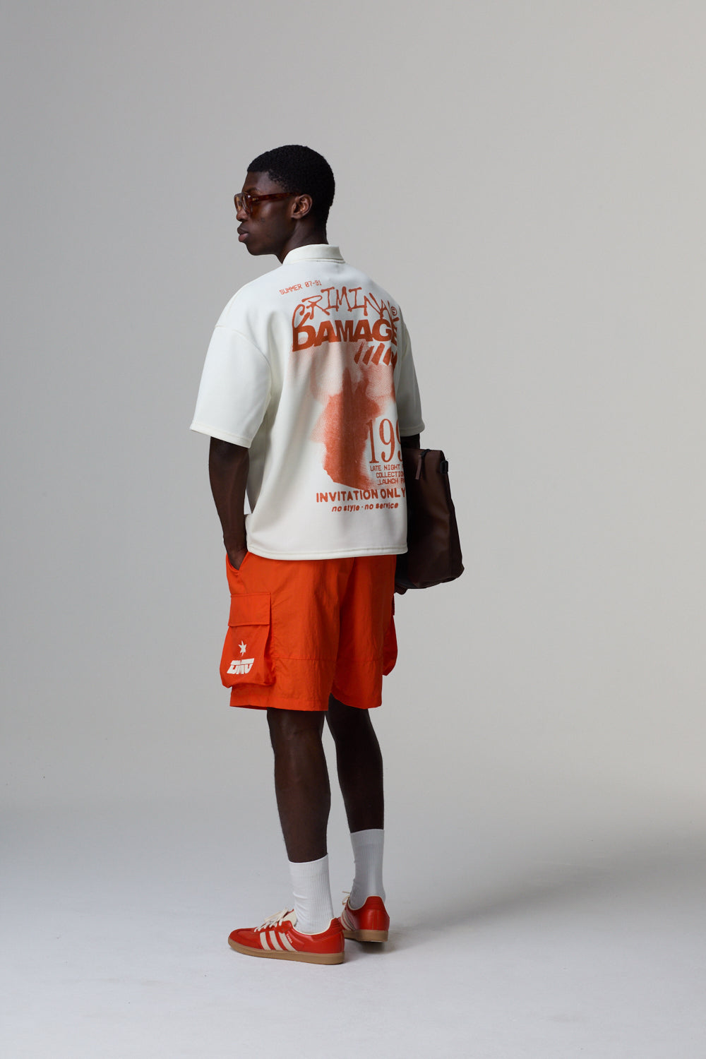 Model wearing a white Criminal Damage polo shirt with graphics and orange shorts on a grey background