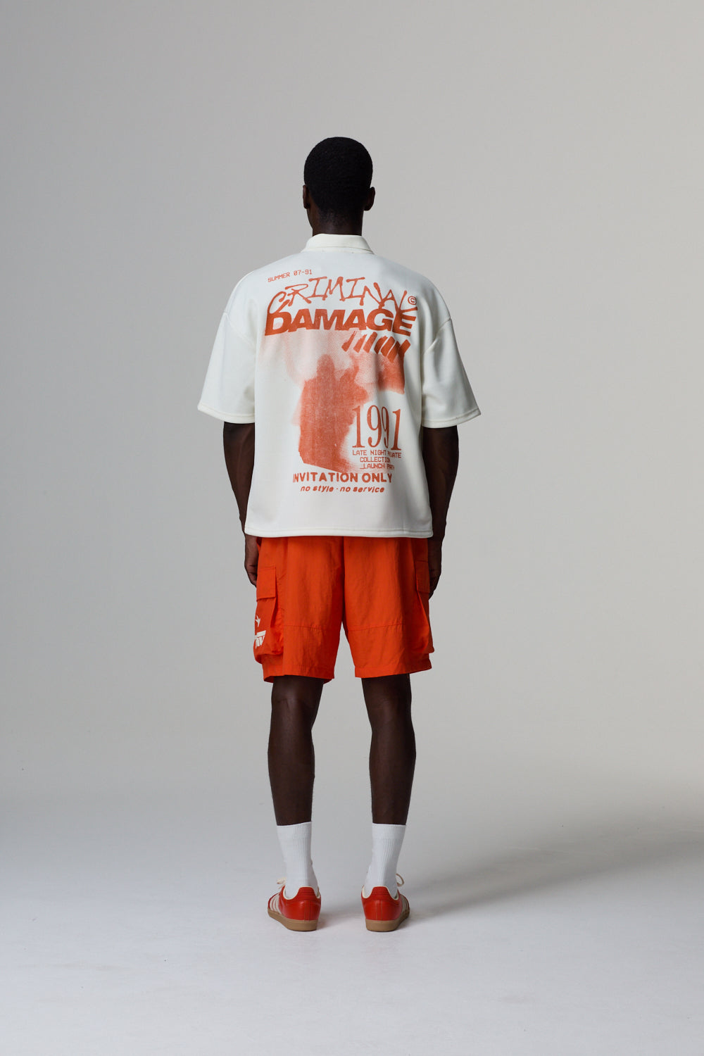 Model wearing a white Criminal Damage polo shirt with text and graphics on the back, paired with orange shorts, standing against a grey background.