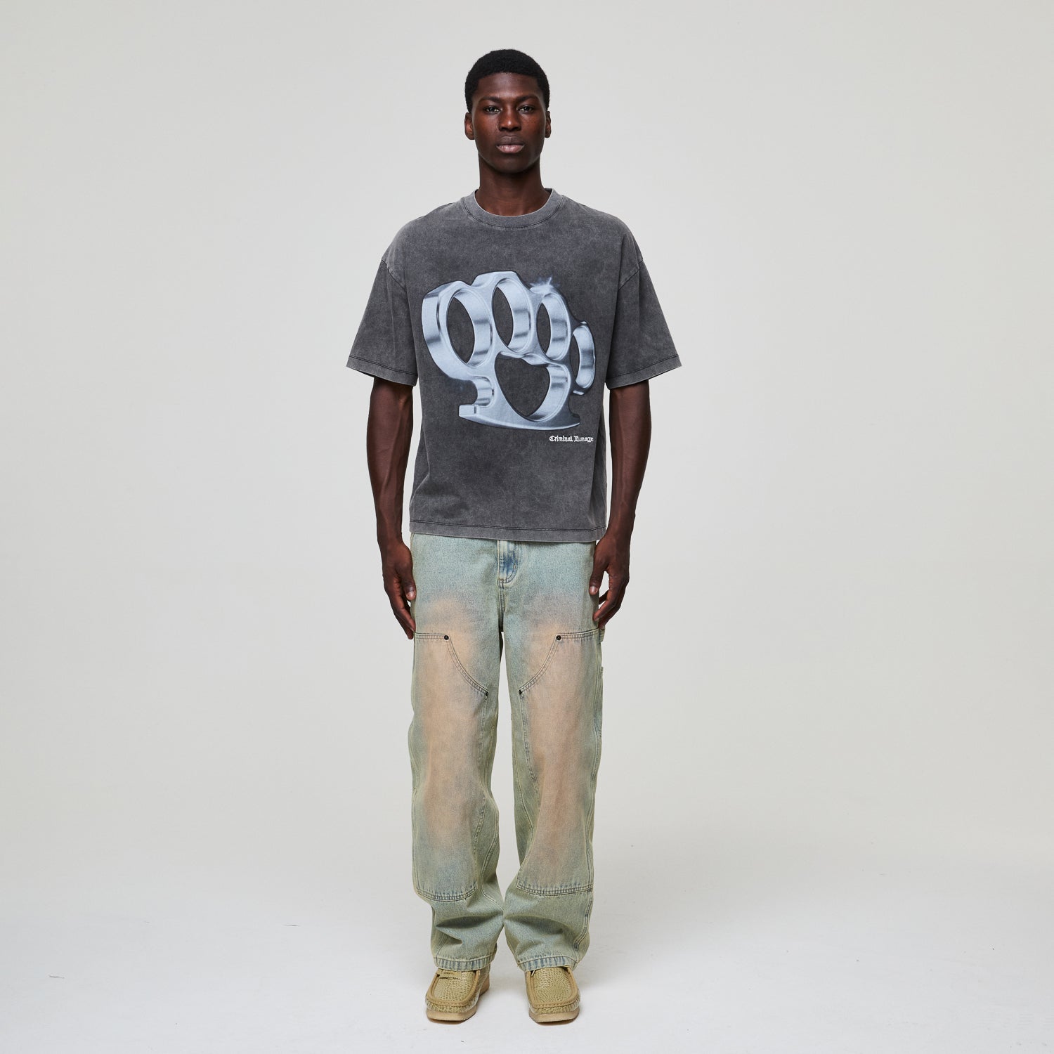 Man wearing a Criminal Damage gray t-shirt with a logo and light blue jeans on a white background