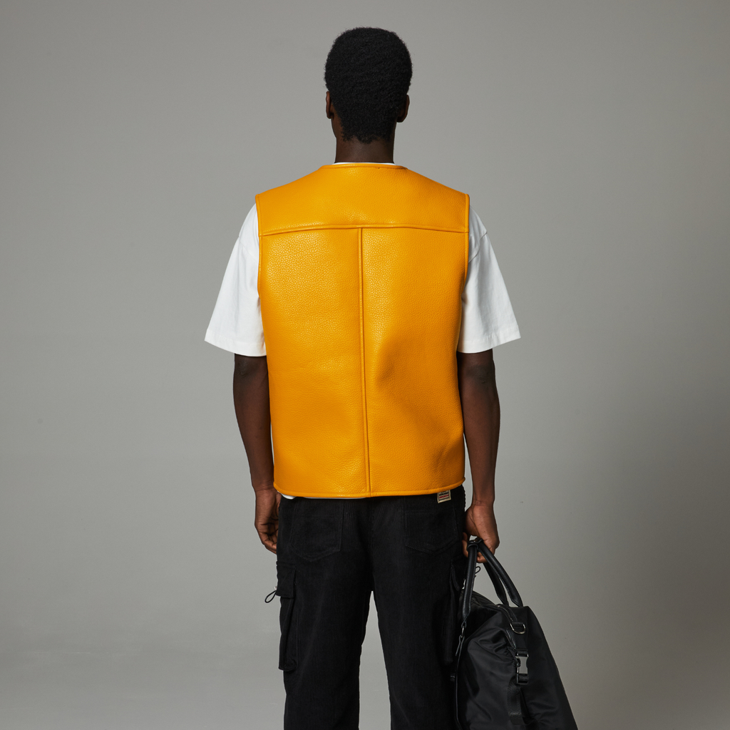 Person wearing a yellow leather vest over a white shirt, holding a black bag on a gray background