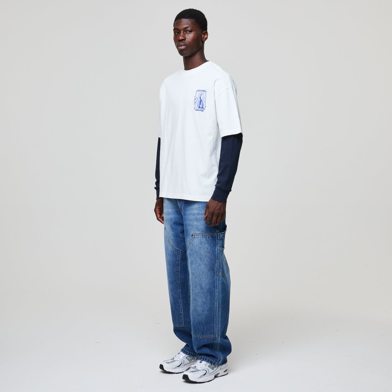 Person wearing a white long-sleeve shirt with a logo and blue jeans on a plain background