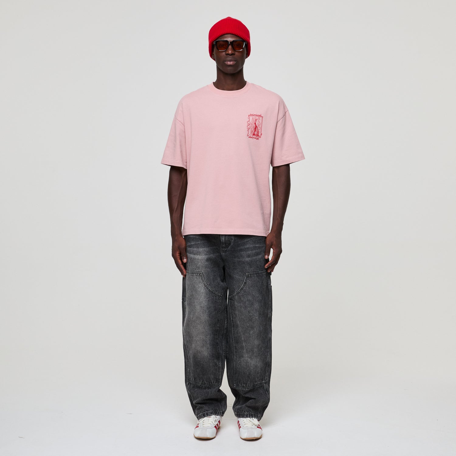 Person wearing a pink t-shirt with a logo, black jeans, and a red cap on a white background