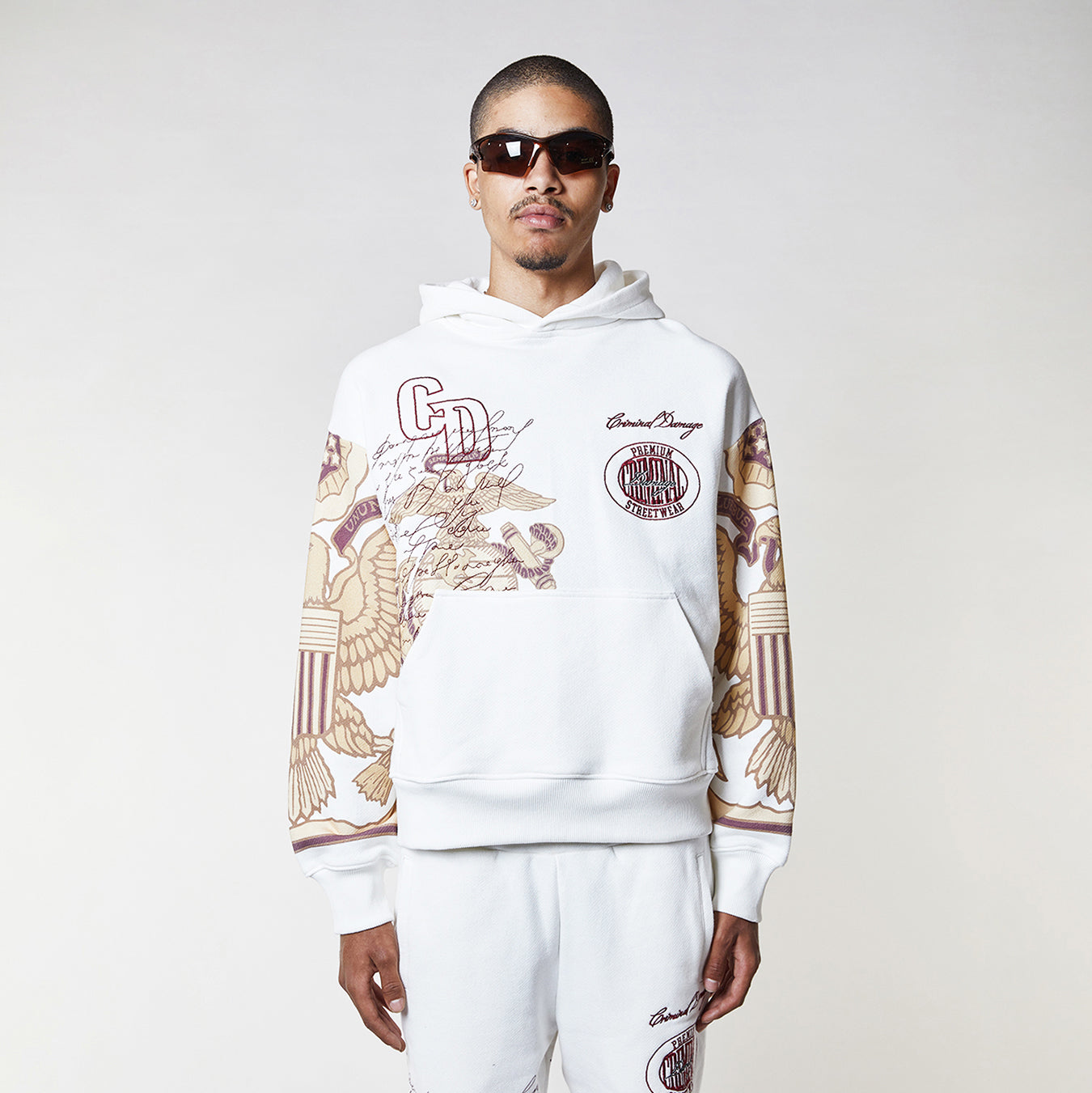 Man wearing a Criminal Damage white hoodie with graphic designs on a plain background