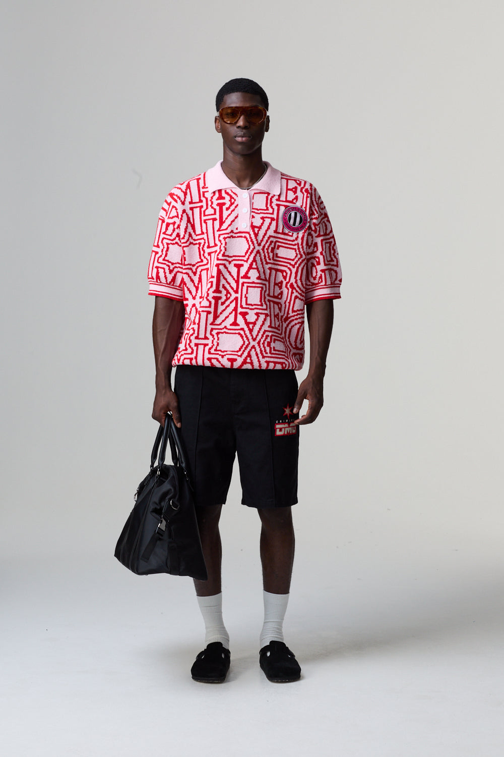 Person wearing a red and white Criminal Damage patterned shirt with black shorts, holding a black bag on a grey background