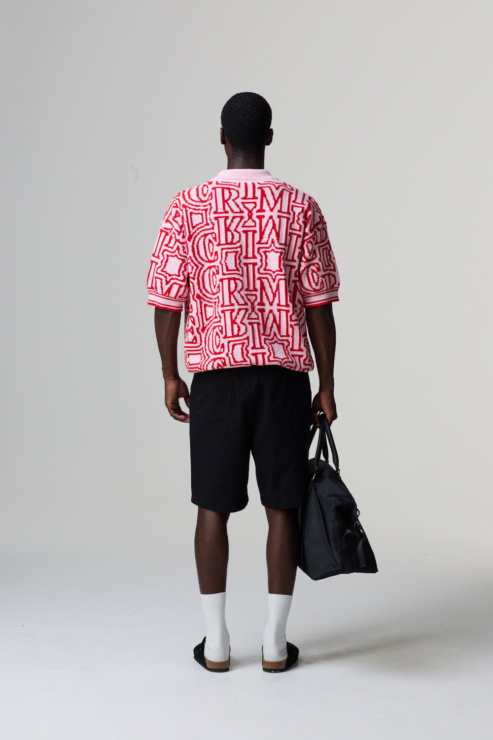 Model wearing a red and white Criminal Damage patterned shirt with black shorts, holding a black bag on a grey background