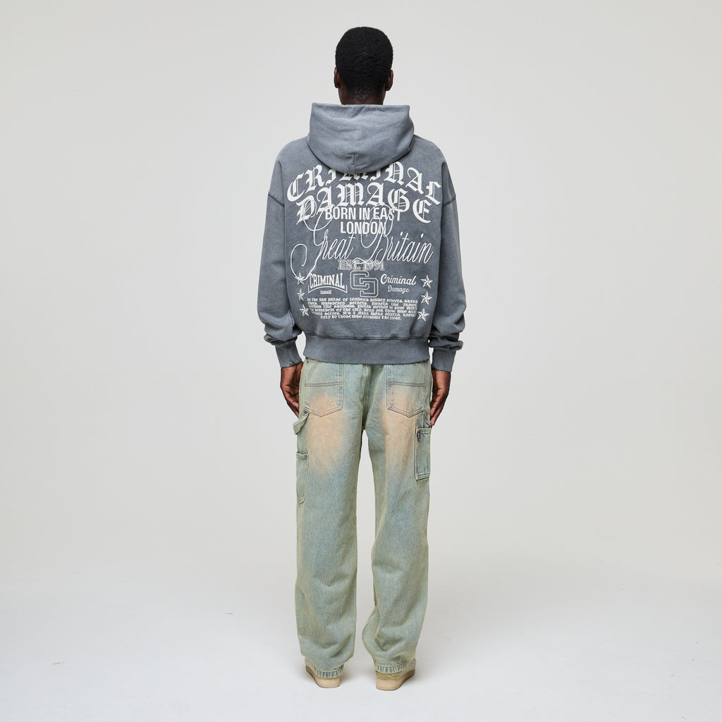 Person wearing a gray Criminal Damage hoodie with text on the back and light blue jeans on a white background