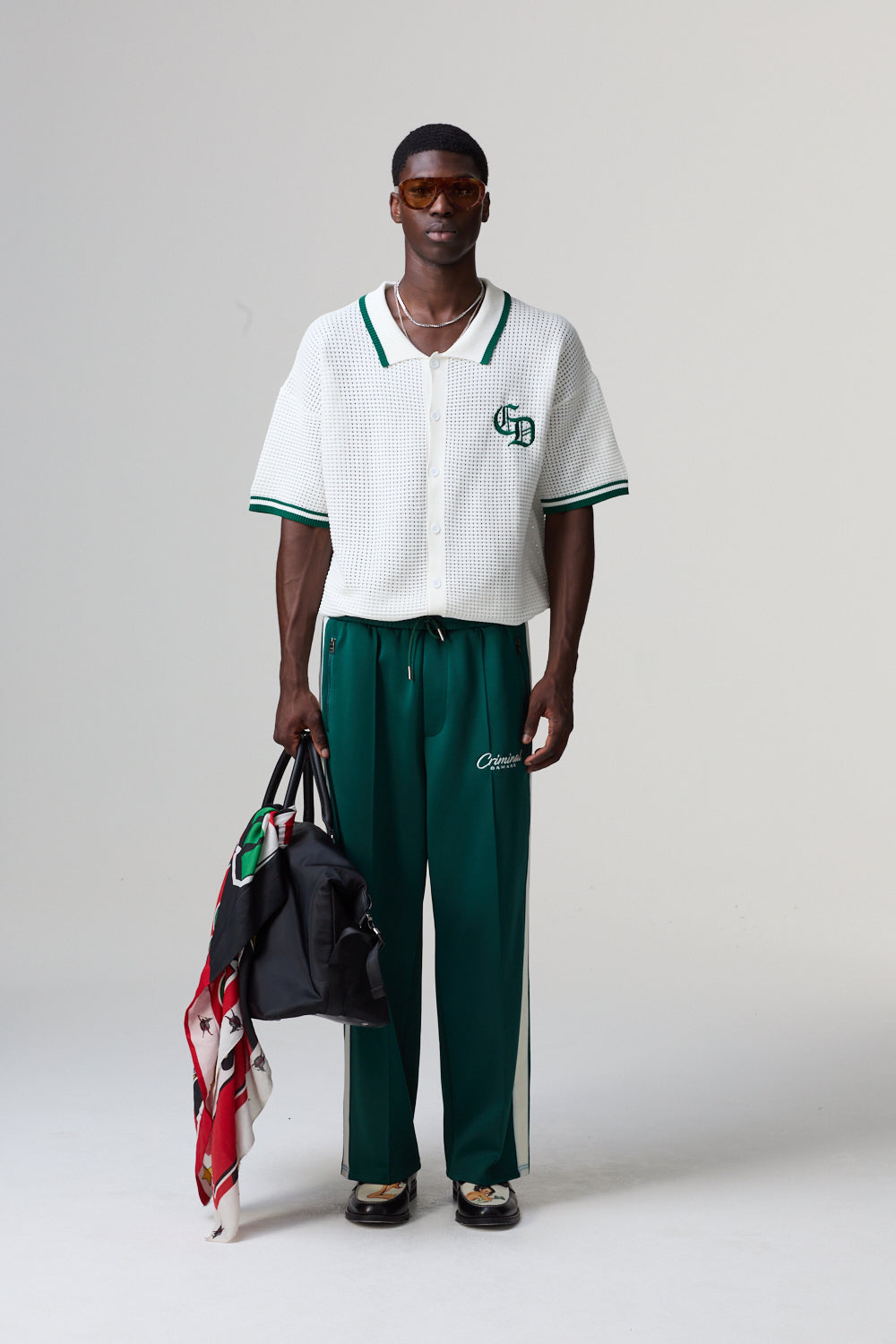 Model wearing a white Criminal Damage polo shirt with green accents and green pants, holding a black bag and colourful scarf.