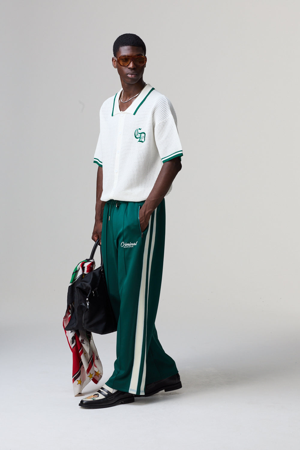 Model wearing a white Criminal Damage polo with green accents and green pants with white stripes, holding a black bag.