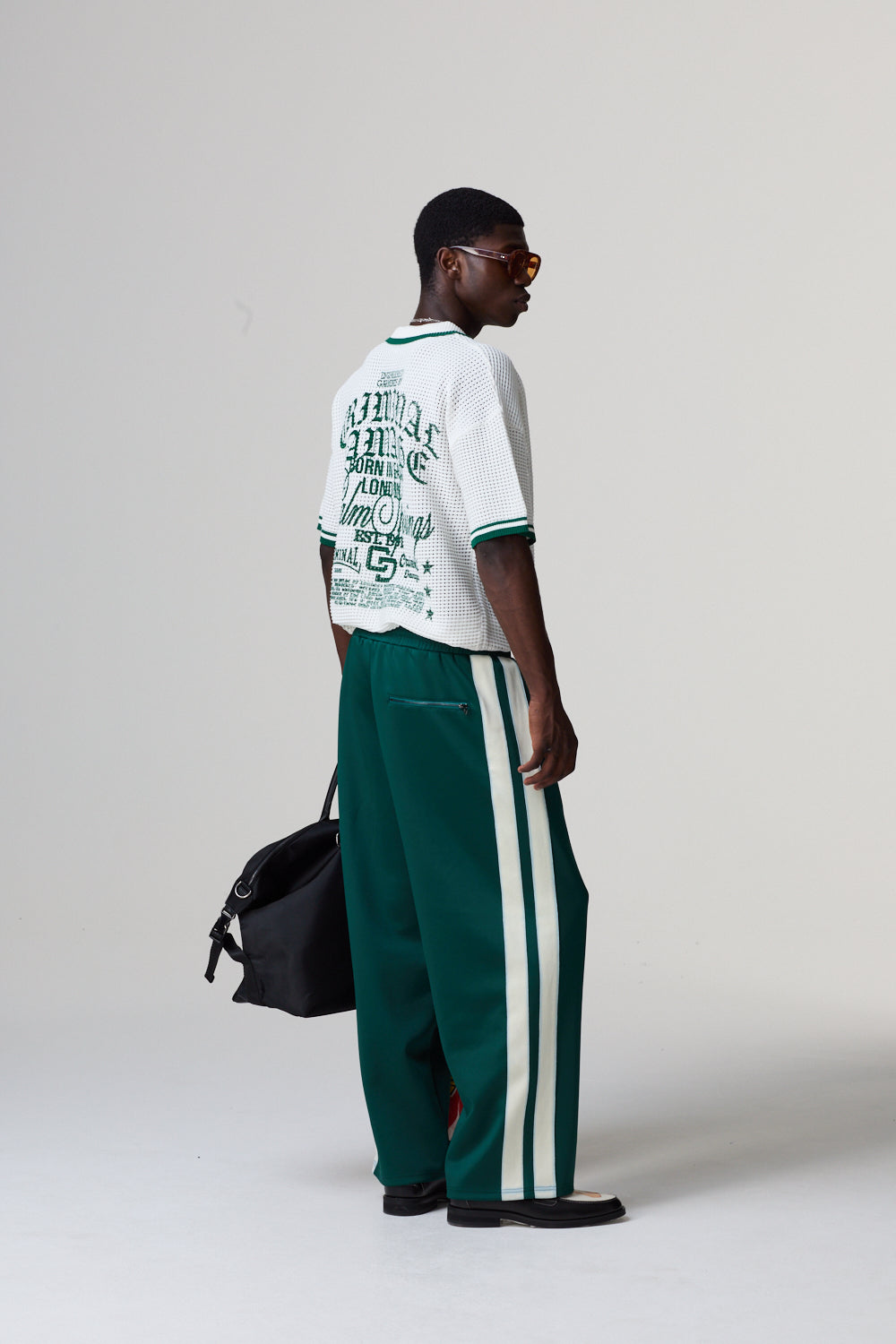 Model wearing a white Criminal Damage t-shirt with text and green pants with white stripes, standing against a grey background.