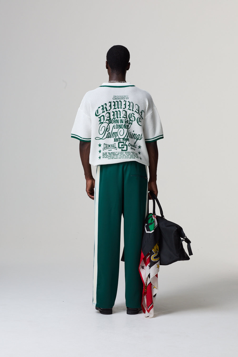 Model wearing a white and green Criminal  Damage outfit with a black bag on a grey background