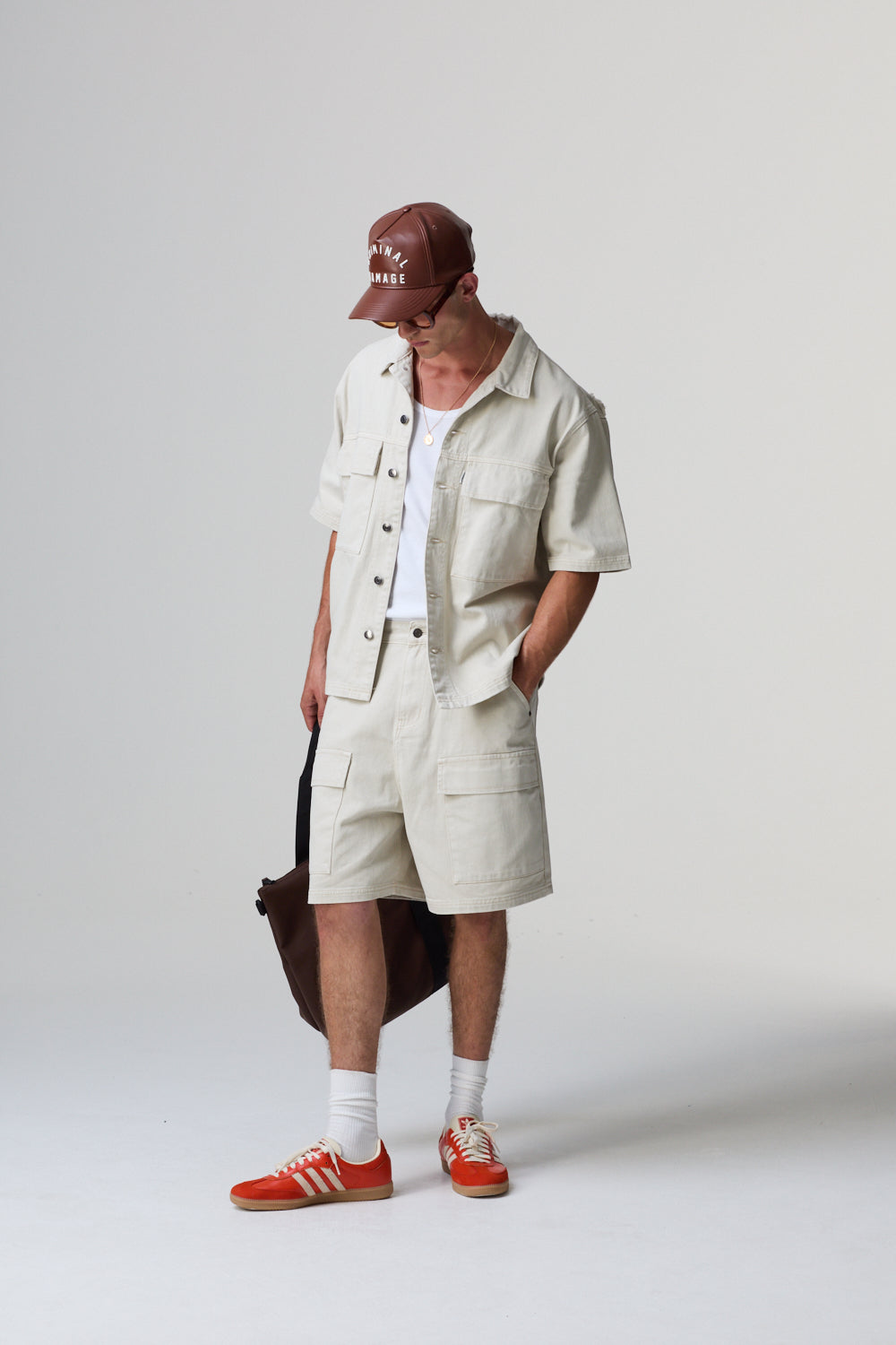 Model wearing a Criminal Damage cotton beige outfit with a cap and red shoes on a grey background