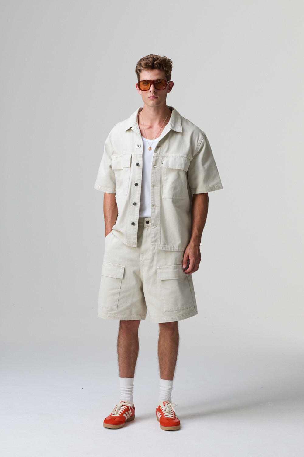 Model wearing Criminal Damage cotton beige outfit with sunglasses on a grey background