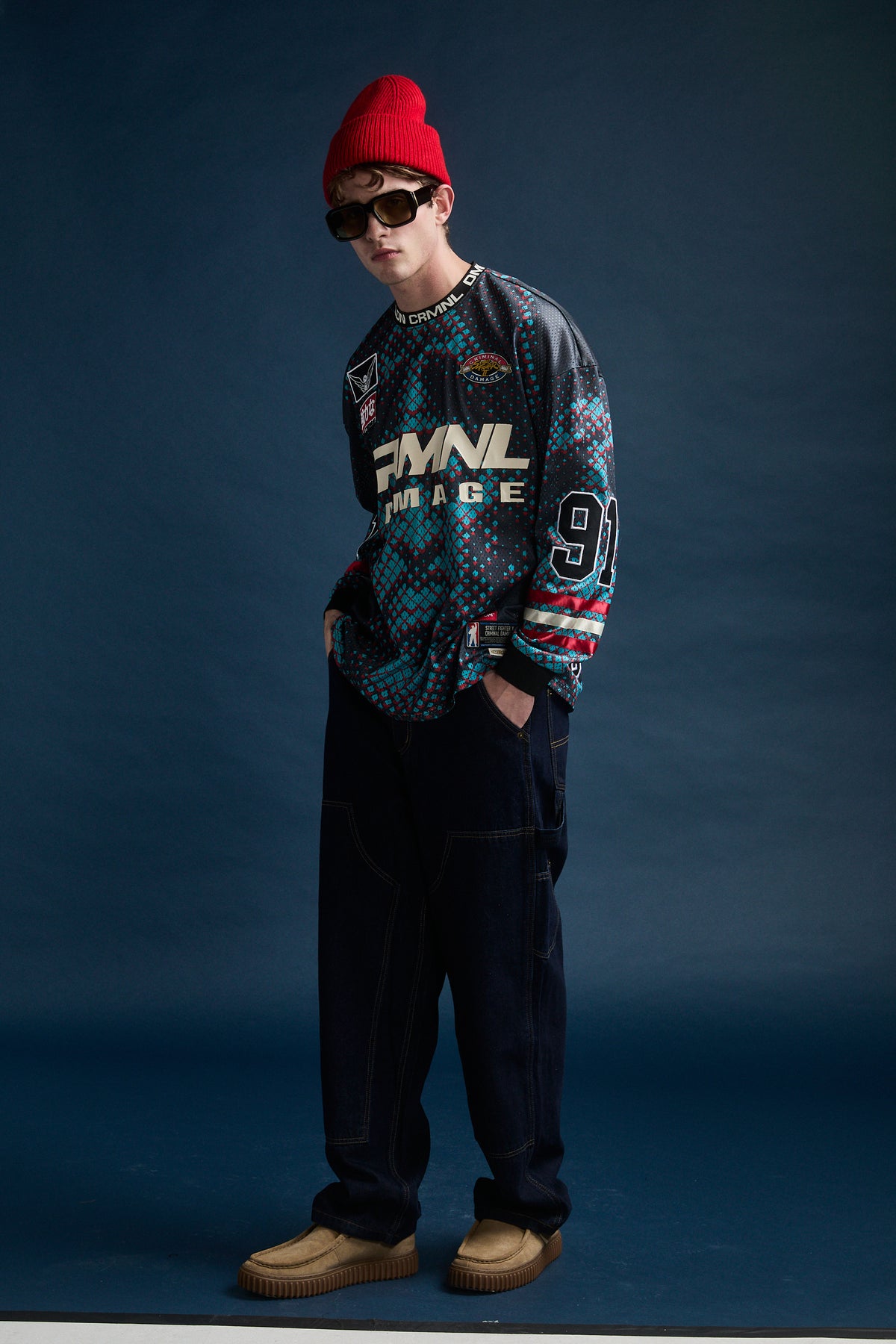 A model wearing a Criminal Damage x Street Fighter with dark pants, a red beanie, sunglasses, and tan slip-on shoes, standing against a dark blue studio backdrop.