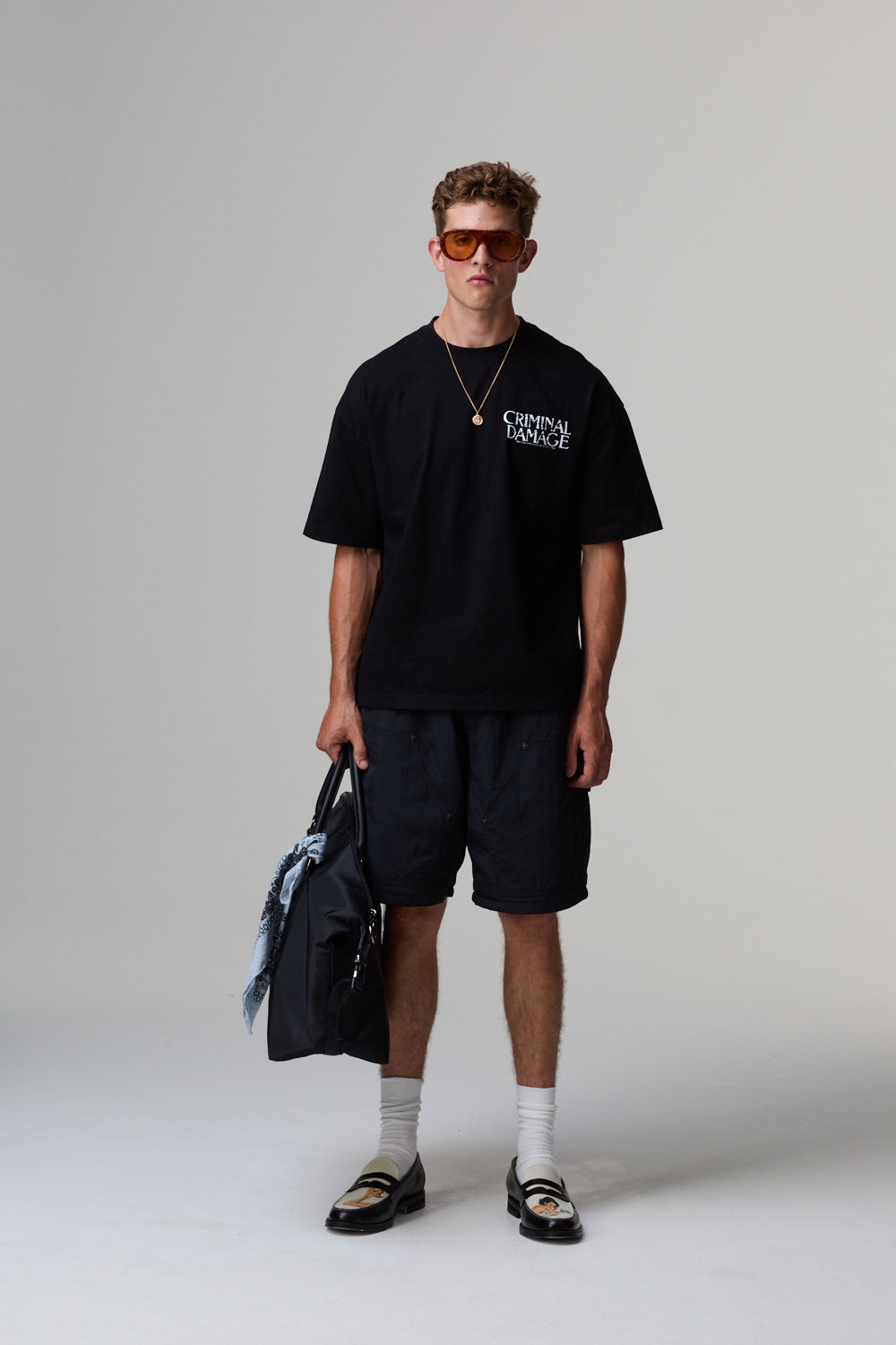 Model wearing a black Criminal Damage t-shirt and shorts with sunglasses, holding a bag on a grey background