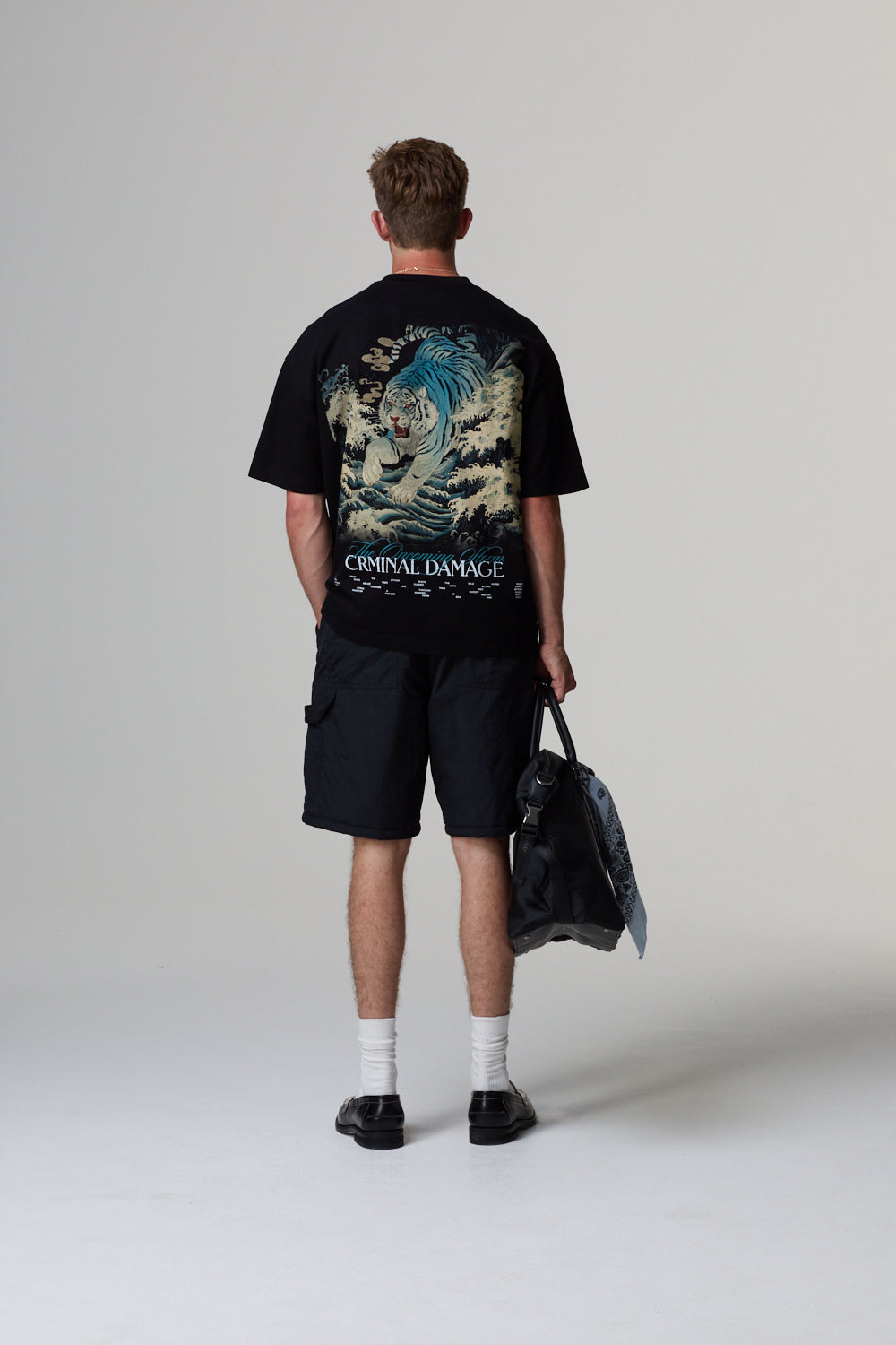 Model wearing a black Criminal Damage t-shirt with a graphic design and text, holding a jacket on a grey background