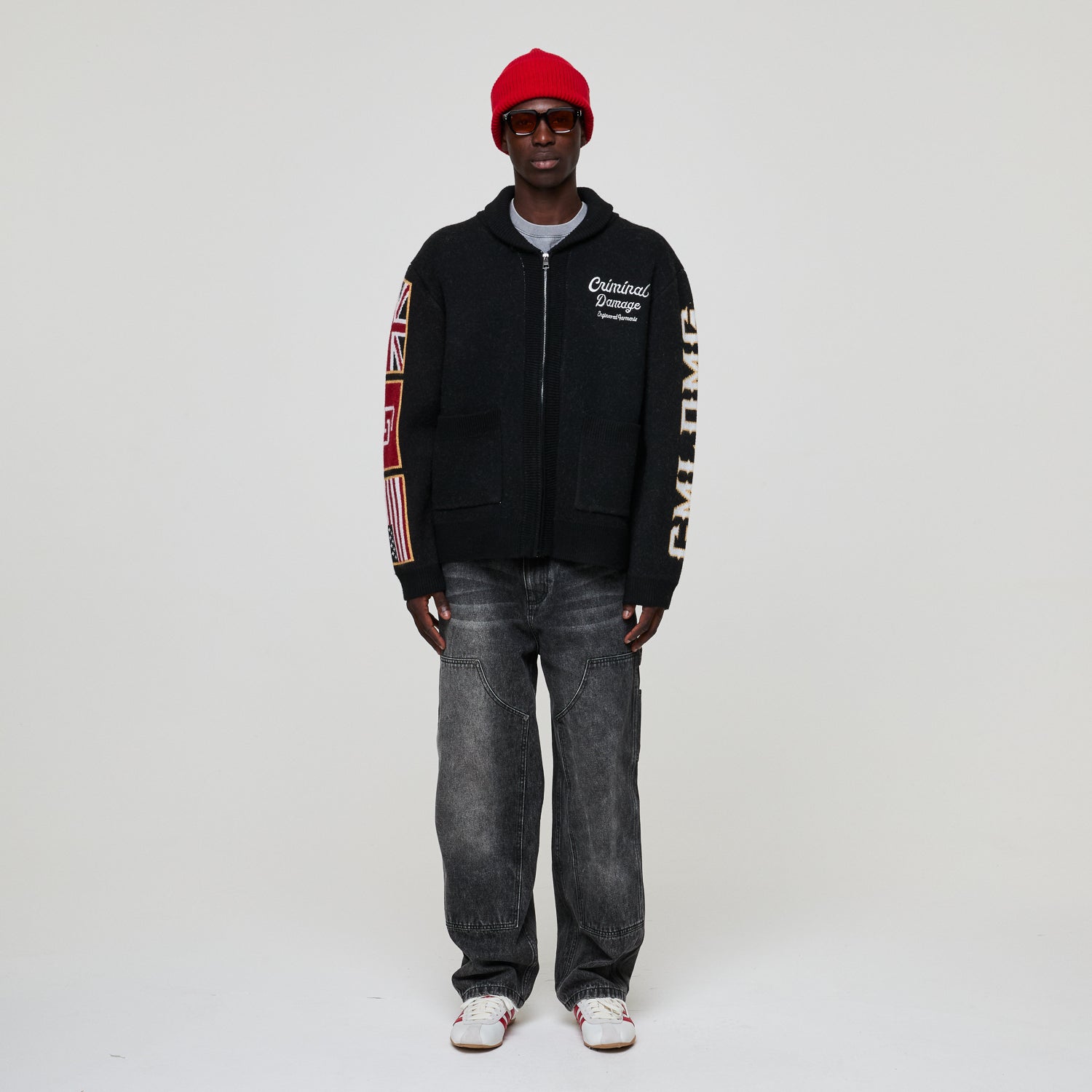 Person wearing a Criminal Damage black hoodie with branding, gray jeans, and a red cap on a white background